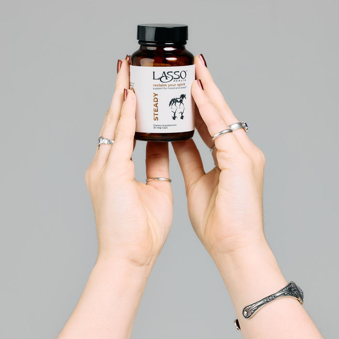 A person's hands holding an amber brown bottle of Lasso Healthy Steady supplement against a solid gray background.