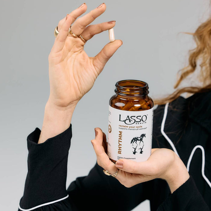Woman holding an open Lasso Health Rhythm supplement bottle and a white capsule.